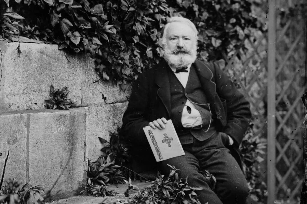 A black and white photograph of a bearded Victor Hugo sitting in the garden